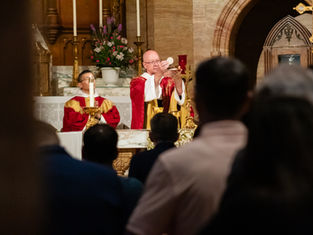 Archbishop elevates communion wafer at altar during Catholic Mass, with candlelit church interior and worshippers in foreground.