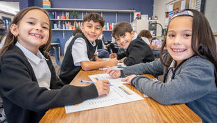 Smiling elementary students at a classroom table with worksheets and markers, learning together in a cheerful classroom.