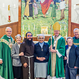 Smiling clergy and lay people pose in a church before an altar, crucifix, and Marian statue, with a bishop holding a staff