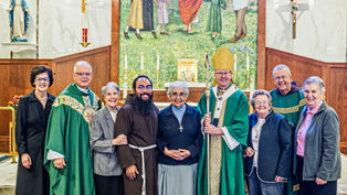 Smiling clergy and lay people pose in a church before an altar, crucifix, and Marian statue, with a bishop holding a staff