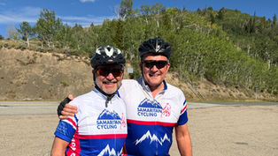 Two smiling cyclists in Samaritan Cycling jerseys and helmets pose on a roadside with forested hills under a blue sky.