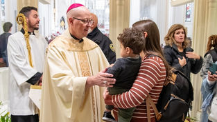 Catholic bishop in white and pink vestments blesses a mother holding a child in a church, with people gathered around them.