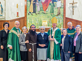 Grupo de religiosos y laicos sonrientes posan en una iglesia, ante altar y crucifijo, con túnicas verdes y mural al fondo.