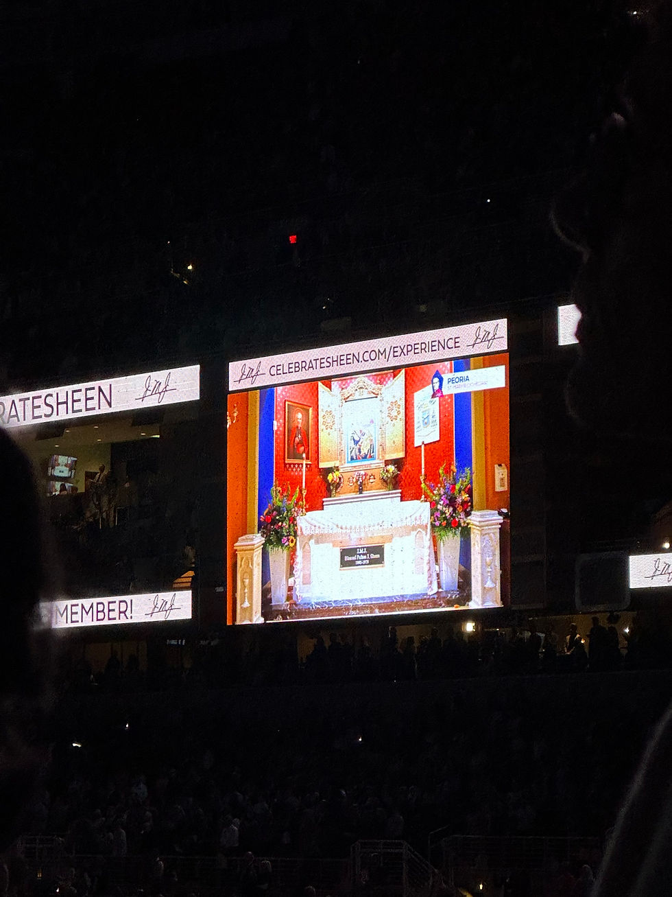 Dark arena with a giant screen showing a colorful altar exhibit; banners read CELEBRATESHEEN.COM/EXPERIENCE and MEMBER!