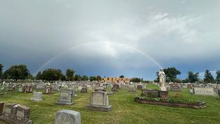 Rainbow over a cemetery with many gravestones, a white statue, and a cloudy sky after rain.