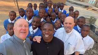 Grupo de escolares sonrientes con dos sacerdotes posando al aire libre en un patio escolar.