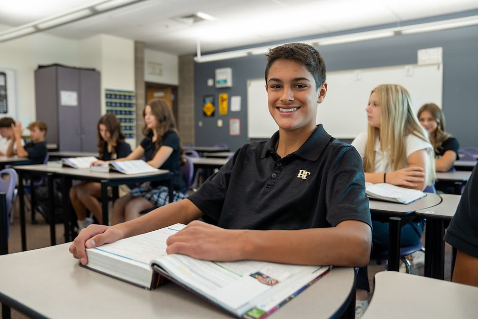 Smiling boy in black polo sits at a classroom desk with an open book, while classmates study behind him near a whiteboard.