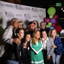 Smiling group poses at a Catholic Charities event backdrop, framed by colorful balloons under an EXIT sign.