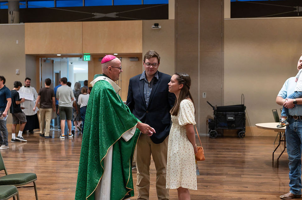 Bishop in green vestments greets a couple in a hall, with people mingling behind them during a church gathering.