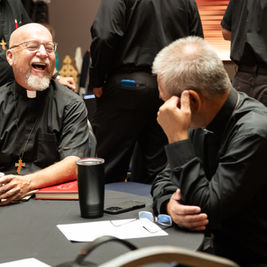 Two clergy in black shirts laugh at a table in a meeting room, with cups, papers, and others standing in the background.