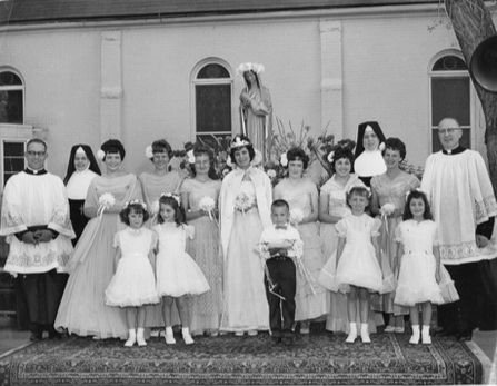 Black-and-white church group portrait of women, children, and priests posed before a statue, with girls in white dresses.