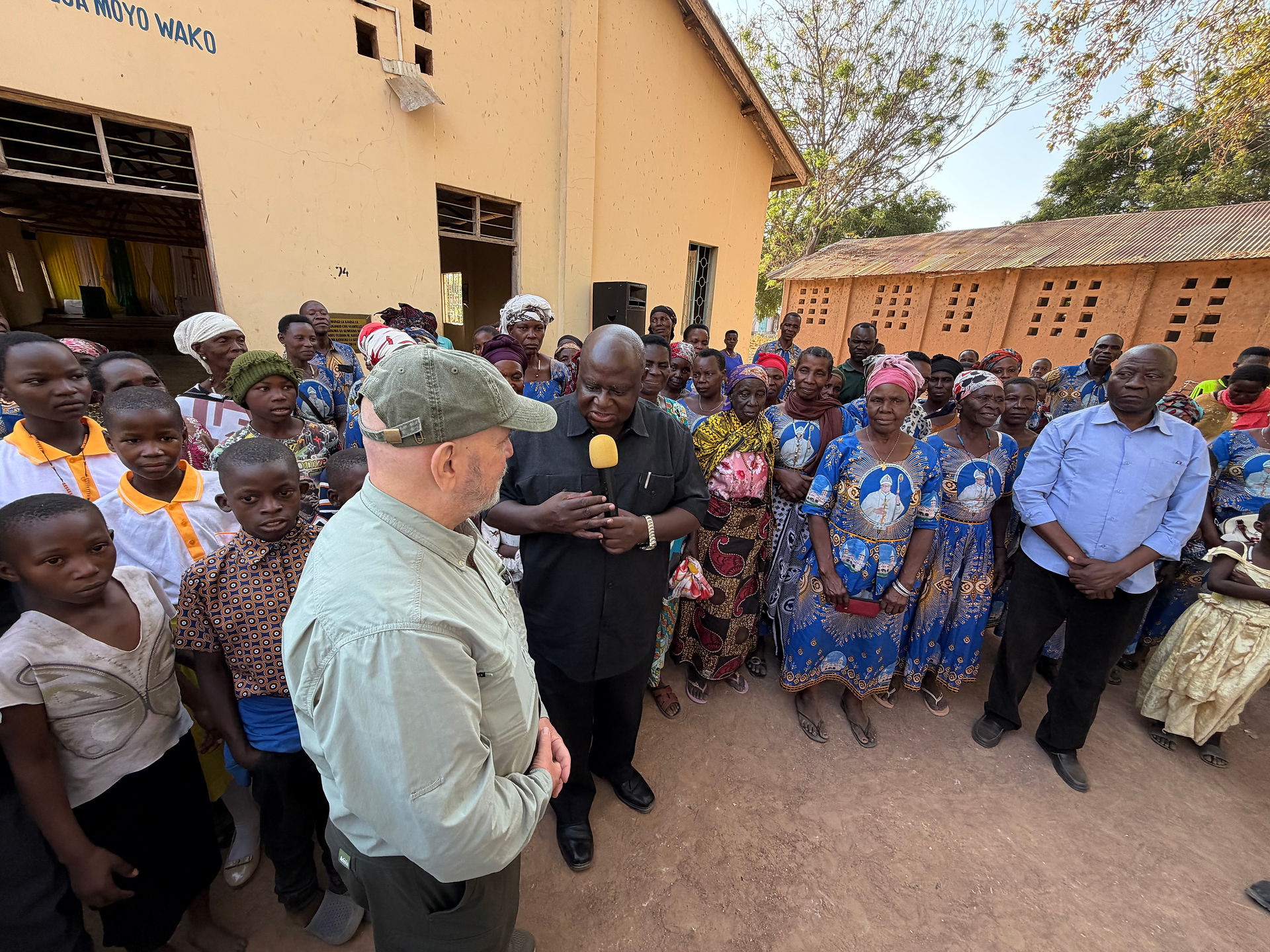 Crowd of villagers outside a beige church or hall listen as a man speaks into a yellow microphone; Moyo Wako text visible.