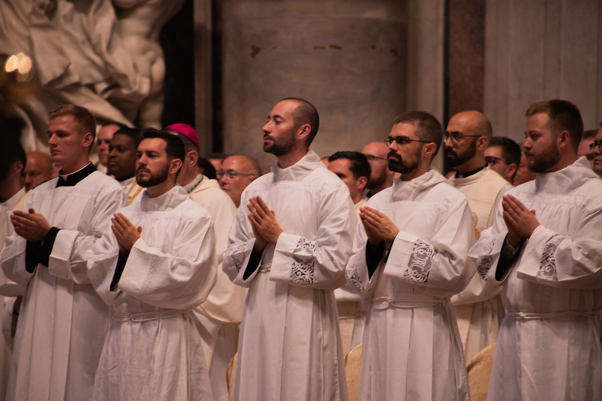Priests in white robes stand praying in a church, hands clasped, with a solemn worshipful mood.