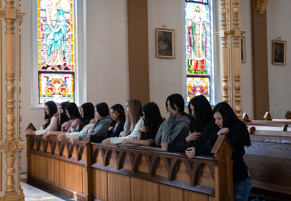 A row of young women sit and pray in a sunlit church, with stained glass windows and wooden pews in a quiet, reverent mood.