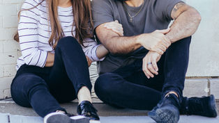 Couple sitting on a sidewalk, leaning together against a white wall, wearing casual black and striped clothes, looking affectionate.