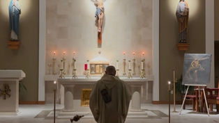 Deacon stands before a candlelit church altar with crucifix and saint statues, facing an easel painting in a quiet chapel.