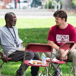 Two men chat and smile on red folding chairs at a park picnic, with plates of food and a water bottle; one shirt says ALWAYS MOVING.