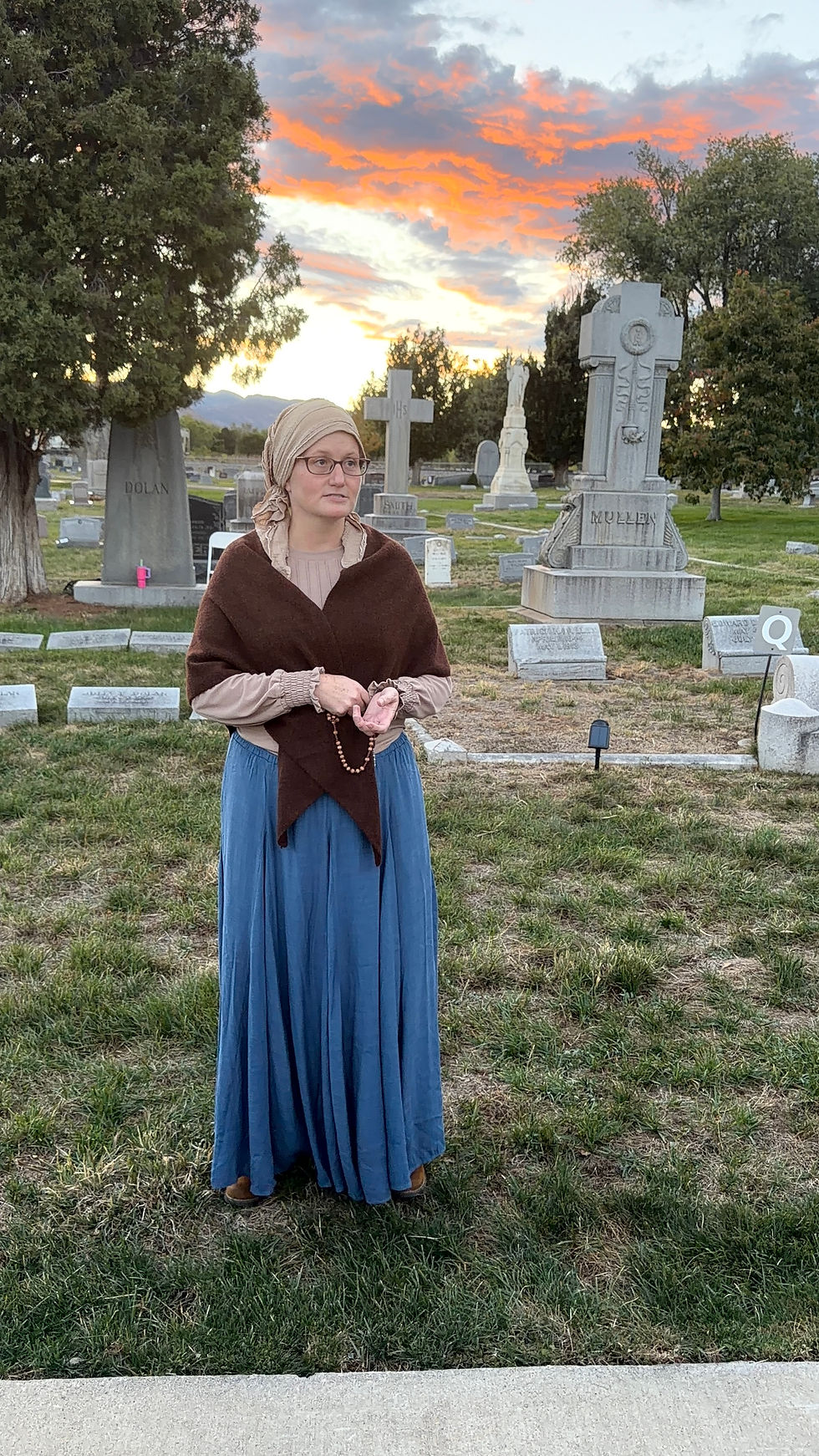 Woman in headscarf and blue dress holds rosary beads in a cemetery at sunset, with orange clouds and gravestones behind her