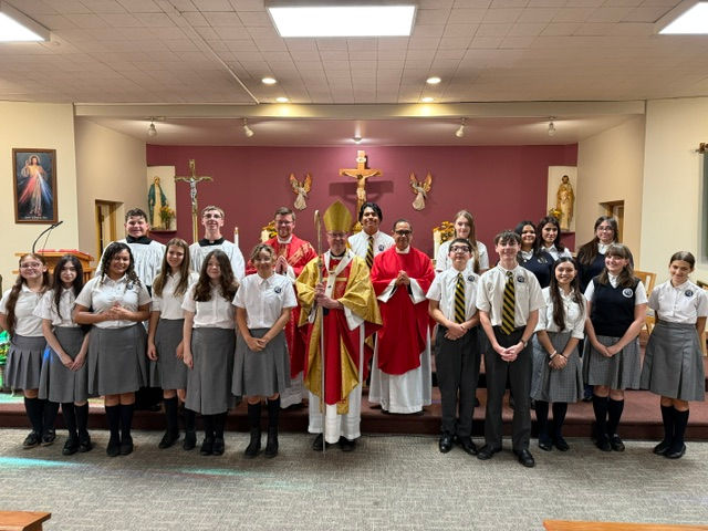 Grupo de estudiantes y clérigos posan en una capilla frente al altar y un crucifijo; colores blanco, rojo y dorado.