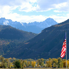 American flag on a pole in front of a mountain valley with autumn trees and snow-dusted peaks under a bright cloudy sky