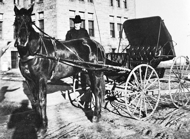 Black-and-white photo of a horse-drawn buggy with a driver in a hat parked beside a building.