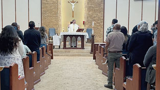 Sacerdote oficia misa en una iglesia; fieles de pie en los bancos, cruz al fondo y vitrales de colores.