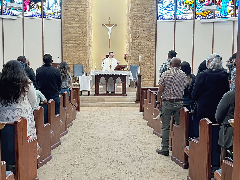 Sacerdote oficia misa en una iglesia; fieles de pie en los bancos, cruz al fondo y vitrales de colores.