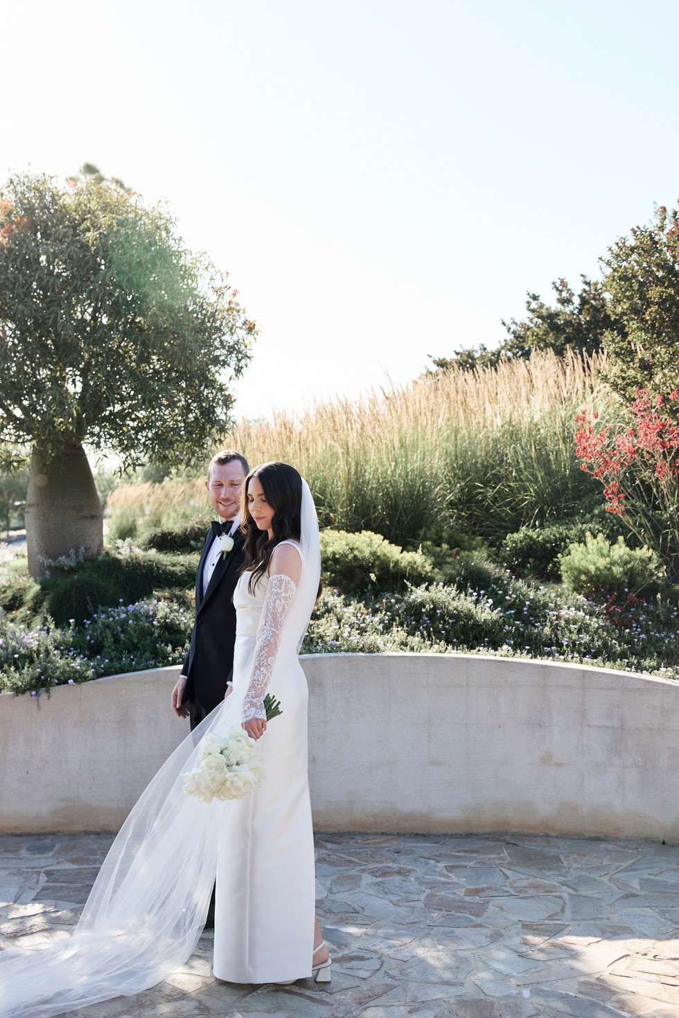 Bride and groom standing in in front of short retaining wall with lush garden in background