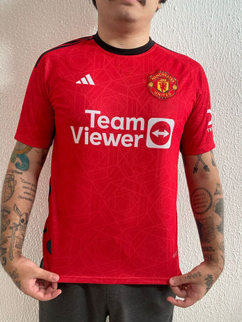 Homem tatuado vestindo camisa vermelha tradicional do Manchester United da temporada 23/24.