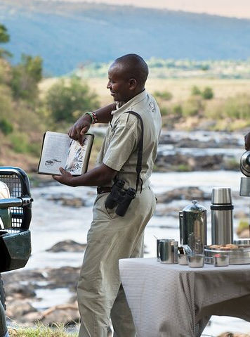 A tourist standing beside the safari jeep, while one guide doing bird interpretation to her by using a field guide book, and another guide preparing a cofee