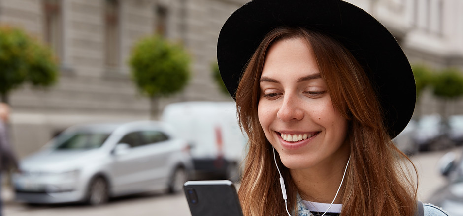 Pleased female in hat, holds cell phone in hand, listens music from playlist, strolls duri