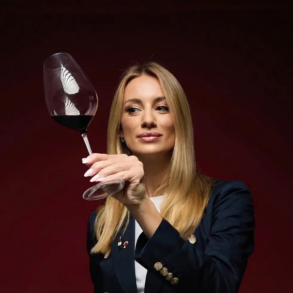 Studio photography of a woman wearing a navy blazer examining a glass of red wine against a deep burgundy background. The scene conveys a sophisticated, expert atmosphere.