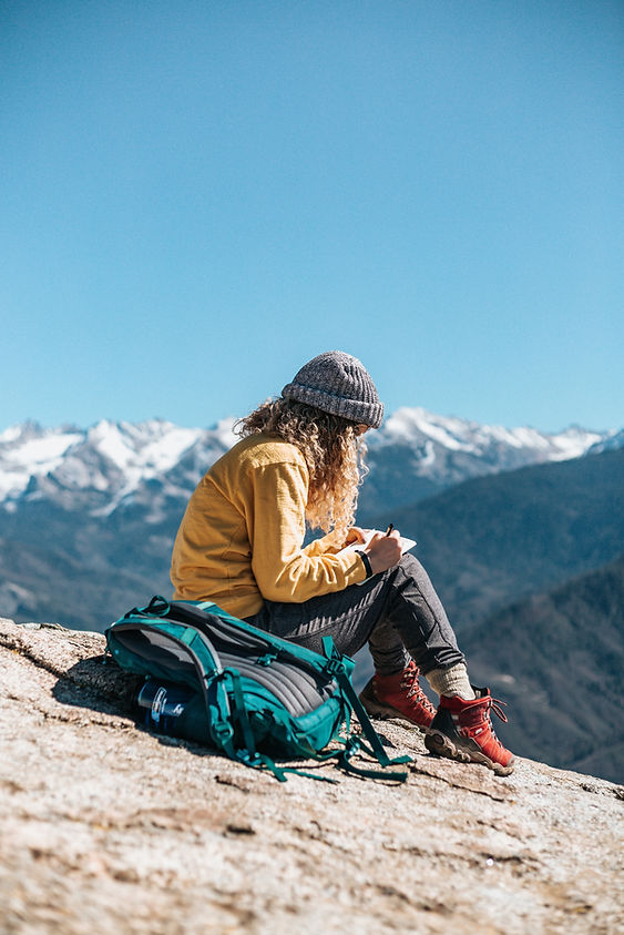 A backpacker sitting on a sloping mountain summit, sketching and taking notes in her travel journal