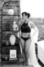 Woman in lingerie poses by a shelf with books and vases against a brick wall. She looks contemplative. Black and white photo.