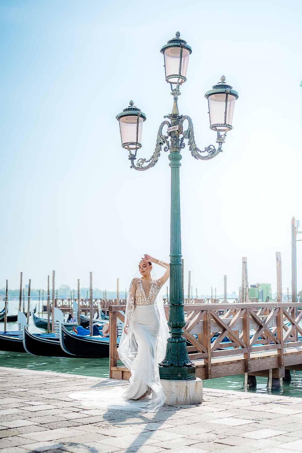 The morning sun graces the outline of a beautiful bride as she poses next to a lamp post on the shores of the Venice canals. Gondolas float in the background. travel wedding photographer, adventure wedding elopement photographer, luxury wedding photographer, Venice, Italy