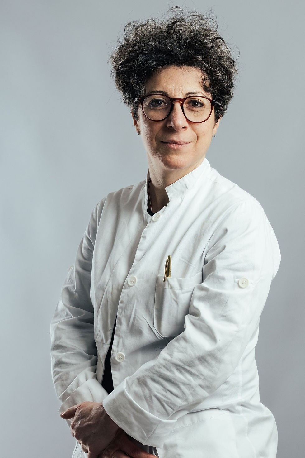 Dott.ssa Manuela De Munari con occhiali e camice bianco su sfondo grigio.