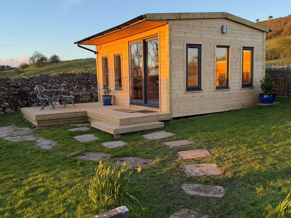 Timber garden room glowing at golden hour, corner windows and bistro set on deck, Cumbria dry-stone wall