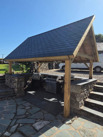Completed outdoor kitchen shelter with slate-effect asymmetric roof and gas BBQ on slate flooring, Cumbria