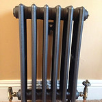 A small, dark gray radiator with five vertical columns stands on a carpeted floor against a beige wall