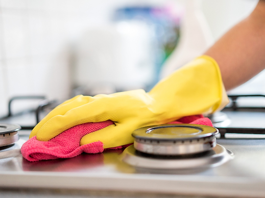 Hand wearing a yellow rubber glove wiping down a stainless steel gas stovetop using a pink microfiber cloth