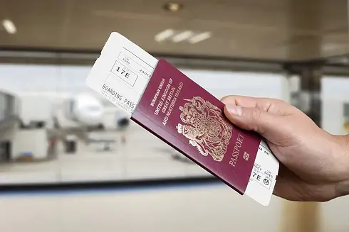 A person holds a maroon UK passport and boarding pass in an airport, with a blurred plane visible through large windows