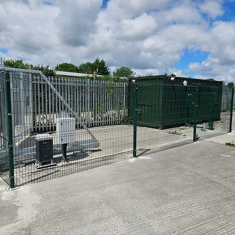 A wide shot showing an enclosed, fenced-off outdoor utility or secure storage area on a concrete ground under a partly cloudy sky