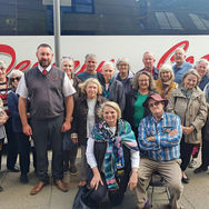 A group of smiling adults, casually dressed, pose in front of a tour bus