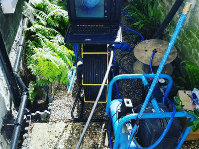 An engineer operating a mobile CCTV drain inspection monitor next to an open manhole in a garden lawn