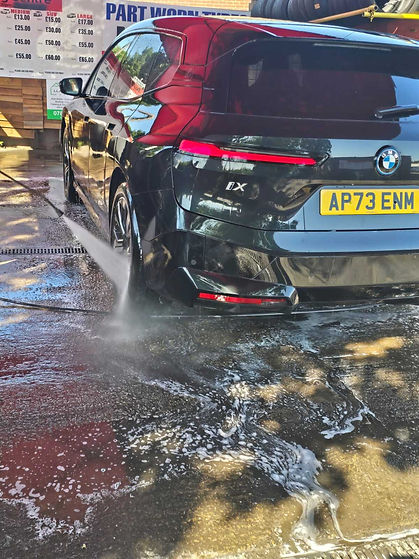 Rear view of a dark grey electric BMW iX SUV being sprayed with a high-pressure water hose at a car wash station