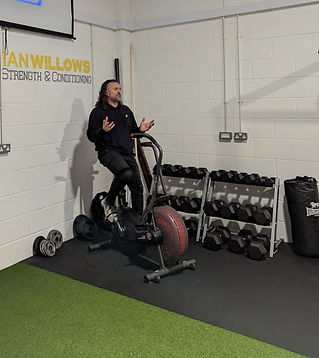 A trainer in a gym explains while standing on a fitness bike, with a screen showing "65%" overhead. Dumbbells and workout equipment surround him