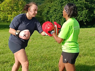 Two women practice boxing outdoors. One in a dark shirt and pink gloves punches red pads held by the other in a neon shirt, with green foliage behind them