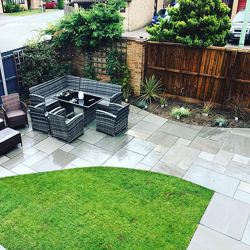 An outdoor patio area featuring grey woven wicker furniture around a dining table, paved stone flooring, and a flower bed along wooden and brick fencing
