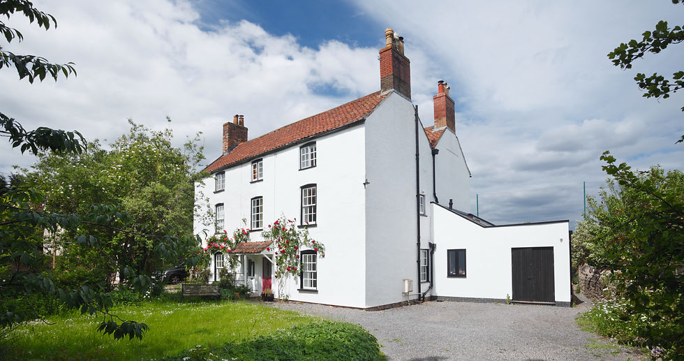 Exterior view of a three-story white countryside house with red roof tiles, chimney pots, climbing red roses over the entrance, and a modern side extension