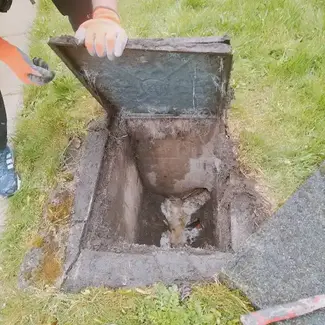 A person wearing orange gloves opens a drain cover in a grassy area, revealing a muddy interior